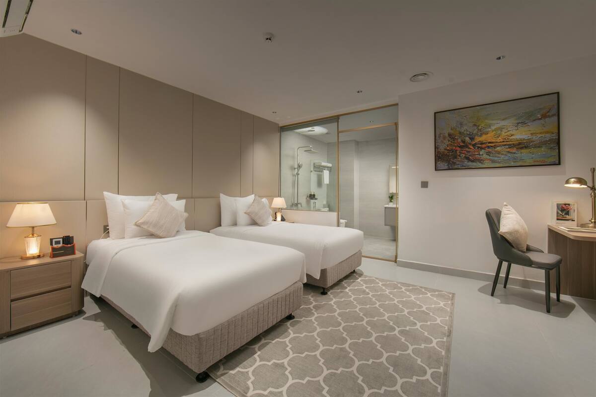 Une chambre d’hôtel moderne avec deux lits, un bureau, une chaise, un grand tableau et une salle de bain visible à travers une porte en verre.