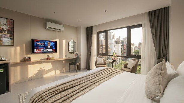Una camera d'albergo moderna con un letto grande, una TV che mostra telegiornali, una scrivania con una sedia e vista sugli edifici dalla finestra.