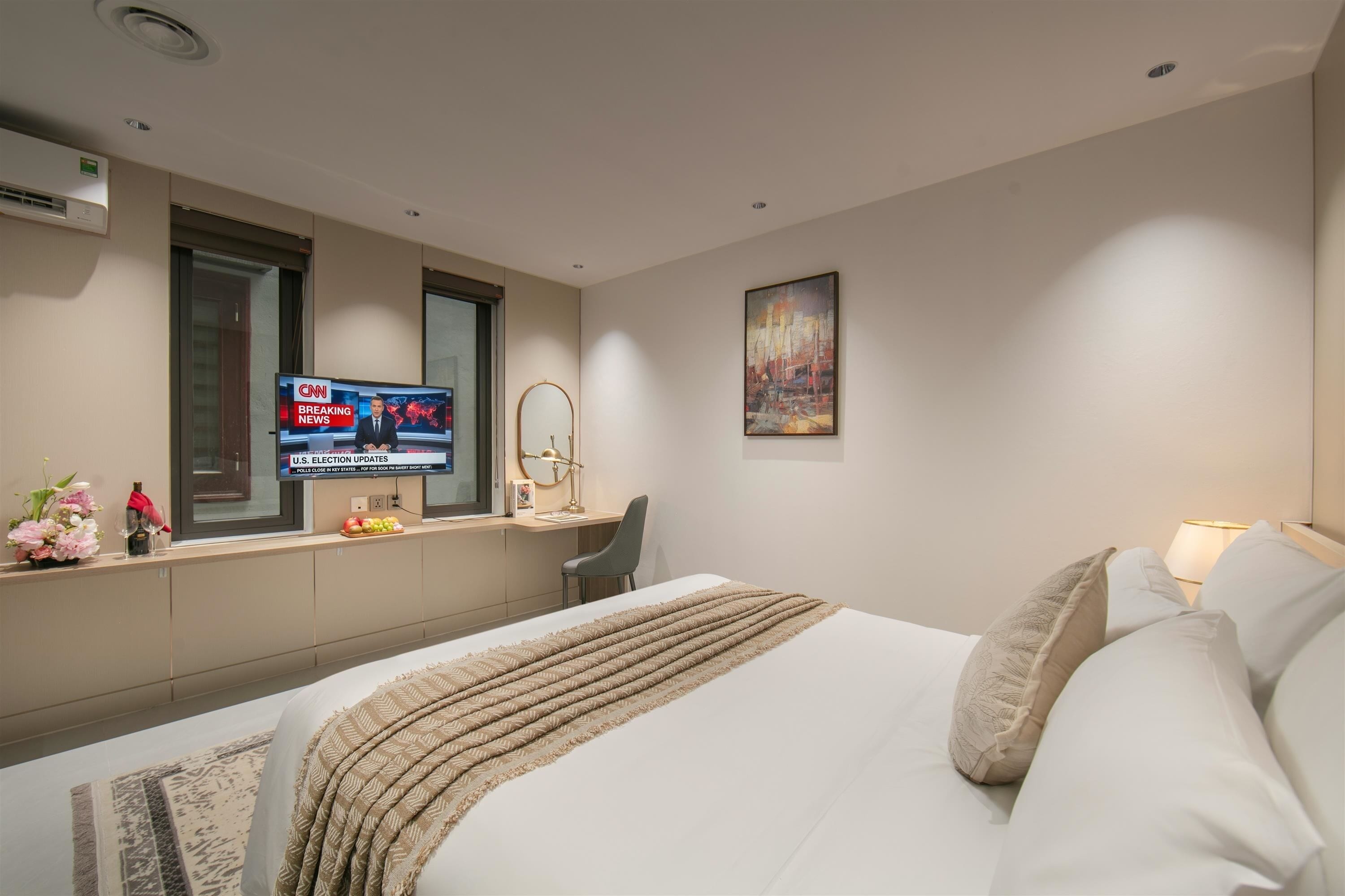 A hotel room with a bed, a TV displaying news, a desk with a chair, a window, and a painting on the wall.