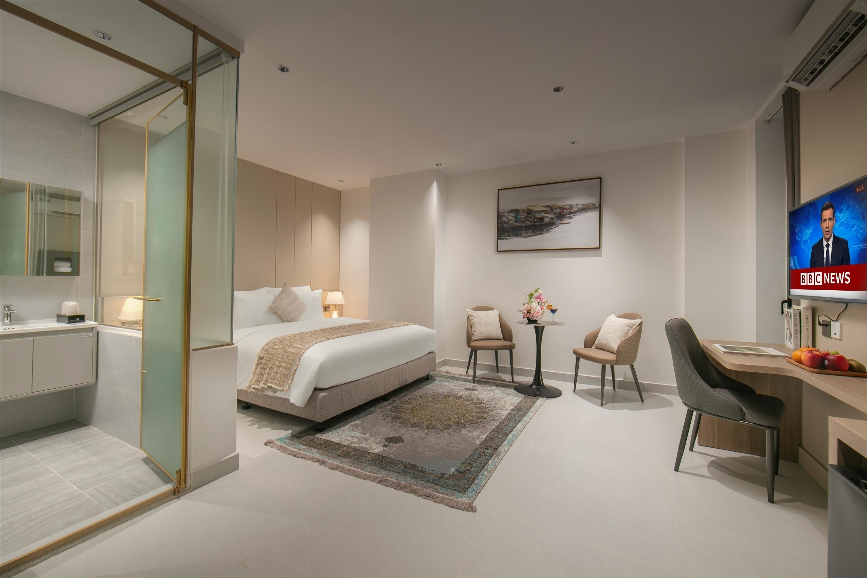 Quarto de hotel moderno com cama, escrivaninha, duas cadeiras, televisão e divisória de vidro.