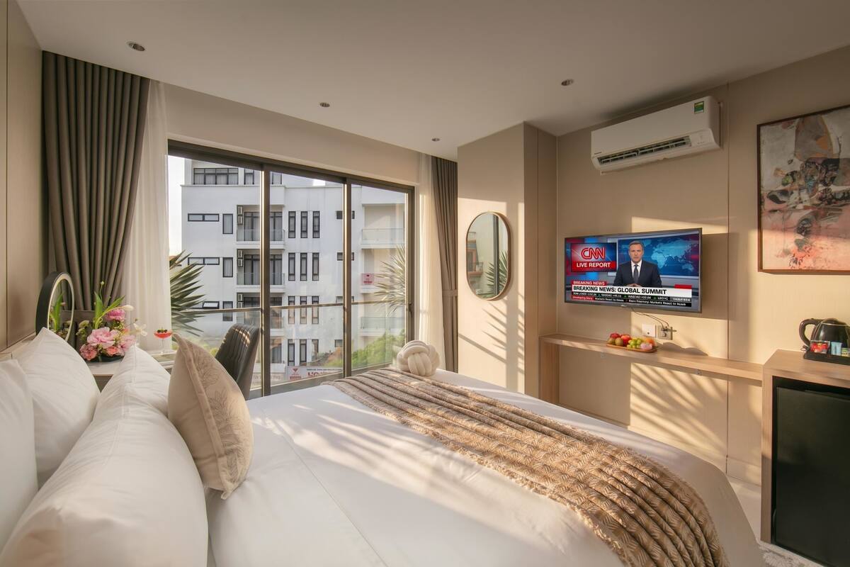Une chambre d’hôtel avec un grand lit, un téléviseur diffusant la chaîne CNN, un balcon avec vue sur les bâtiments et un appareil de climatisation.