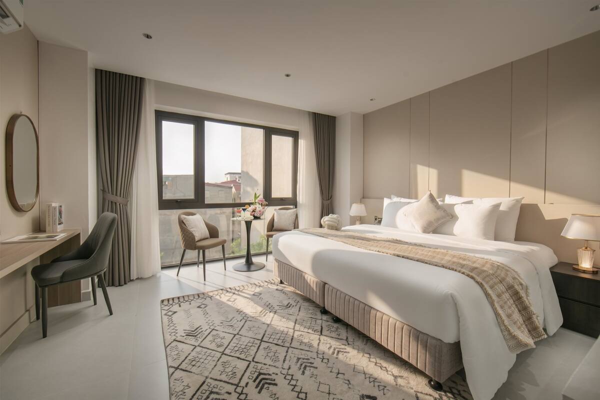 Une chambre d’hôtel moderne avec un grand lit, un bureau, une chaise, une petite table agrémentée de fleurs et une vue sur la ville.