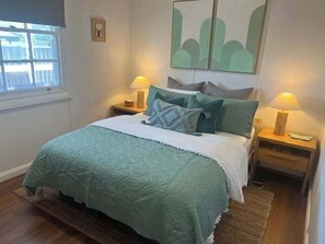 Una cama bien tendida con una manta verde, dos lámparas de noche y un cuadro en la pared.