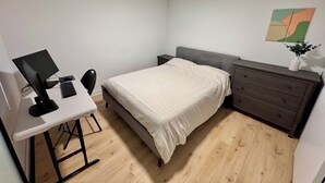 Ein Schlafzimmer mit Bett, Schreibtisch mit Computer, Kommode und einem Wandbild.