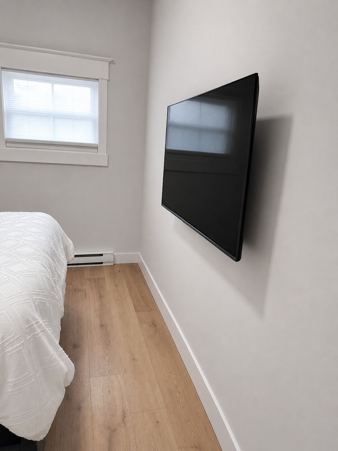Une chambre à coucher avec un lit, un téléviseur fixé au mur, une fenêtre et un plancher en bois.