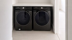 Una sala de lavandería con dos lavadoras de carga frontal, techo blanco y una puerta que da a otra habitación.