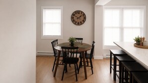 Un comedor con una mesa redonda, cuatro sillas, un reloj de pared y dos ventanas con persianas.