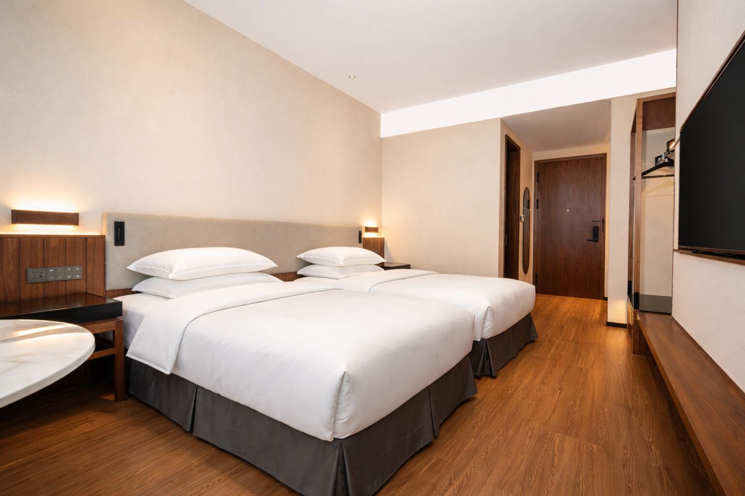 A hotel room with two beds, a desk, a TV, and a wooden floor.