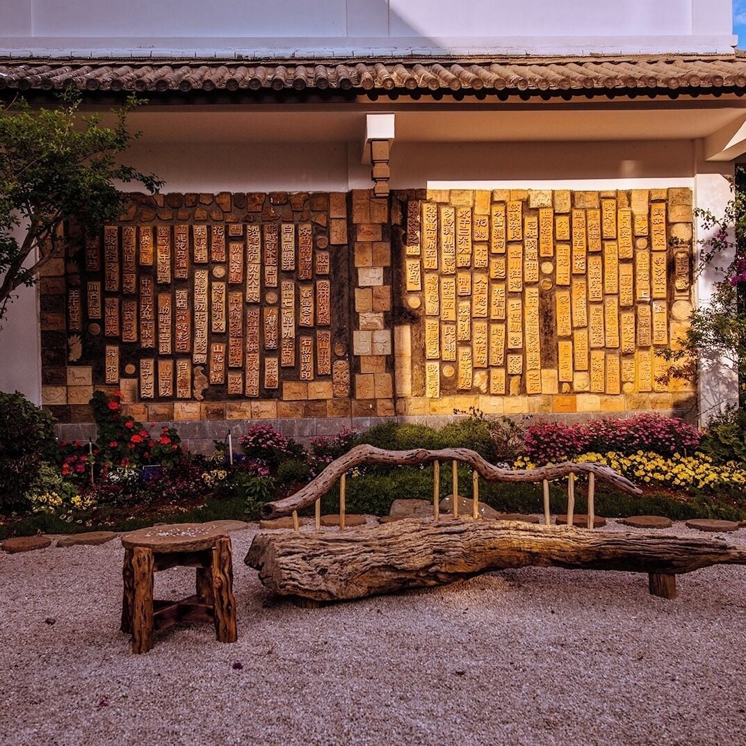 A courtyard with a stone wall featuring carved inscriptions, a wooden bench, and a rustic log structure.