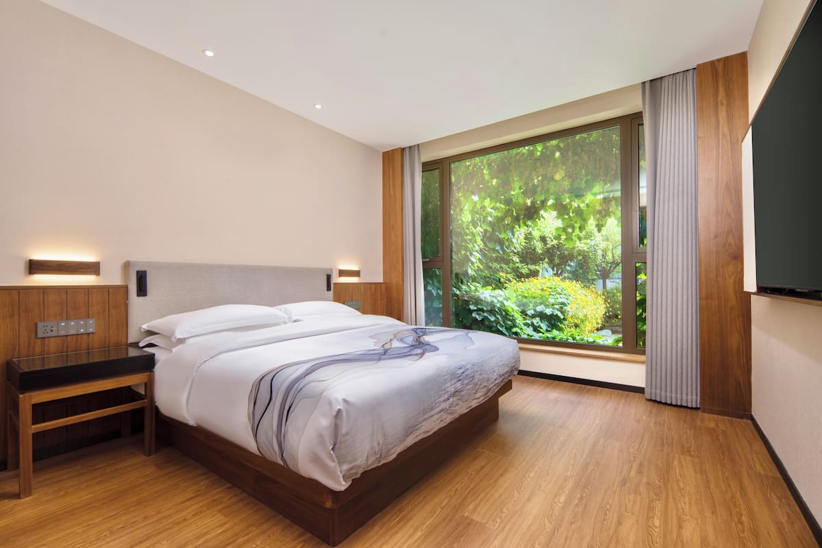 Een moderne slaapkamer met een groot raam, een houten bed, een nachtkastje en een aan de muur bevestigde televisie.