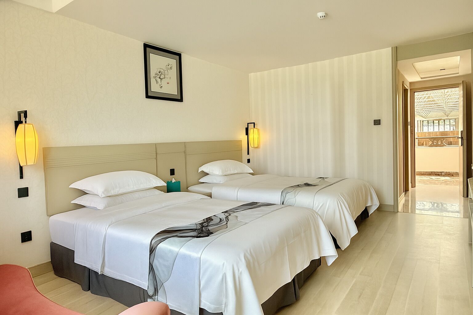 Camera d'albergo con tre letti, un comodino con una lampada, un quadro appeso alla parete e vista sull'esterno.