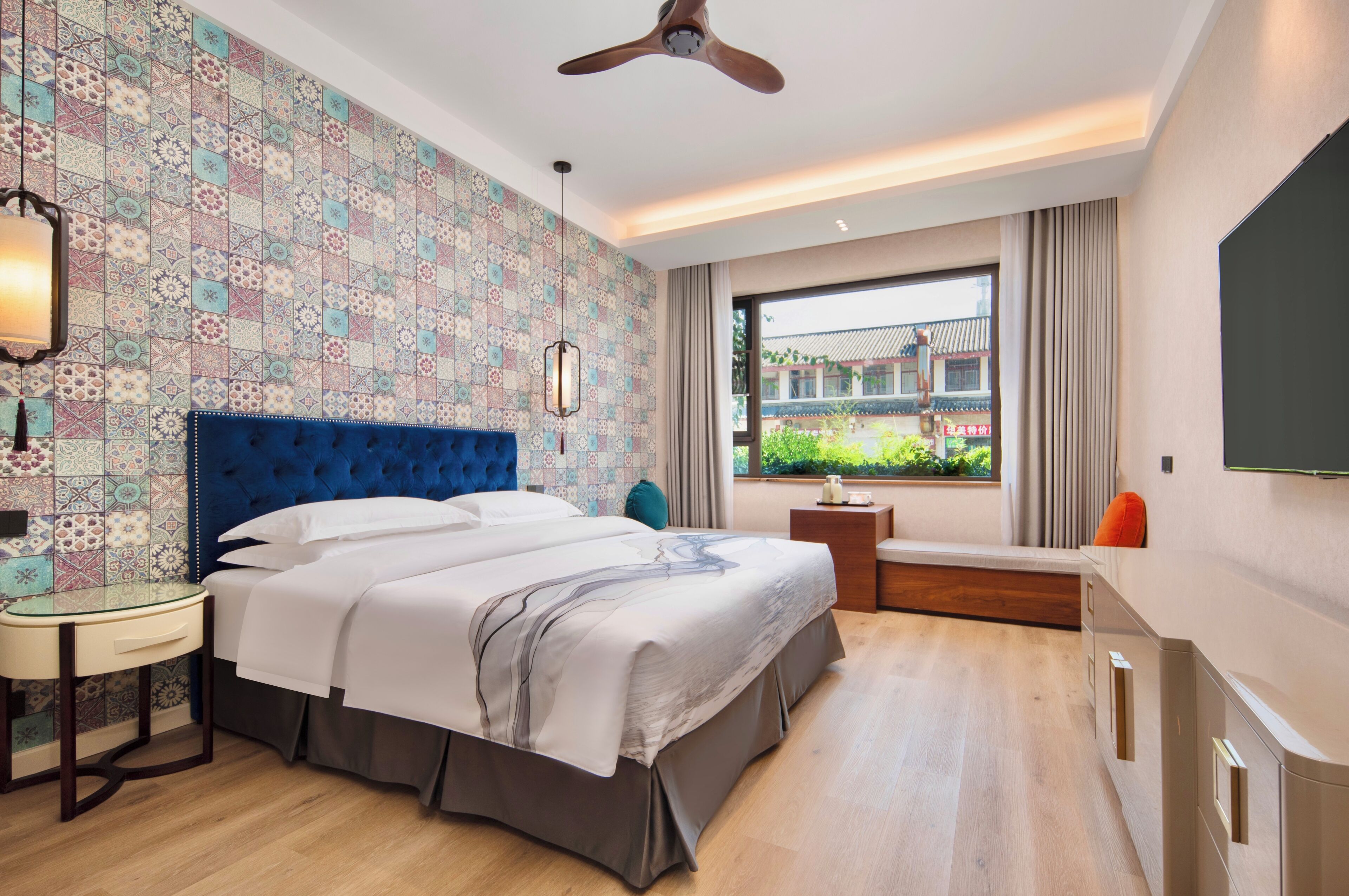 Ein Hotelzimmer mit einem großen Bett, einem Deckenventilator, einem Fernseher, einem Fenster mit Blick auf Grünflächen und traditionelle Architektur sowie einer Wand mit Muster.