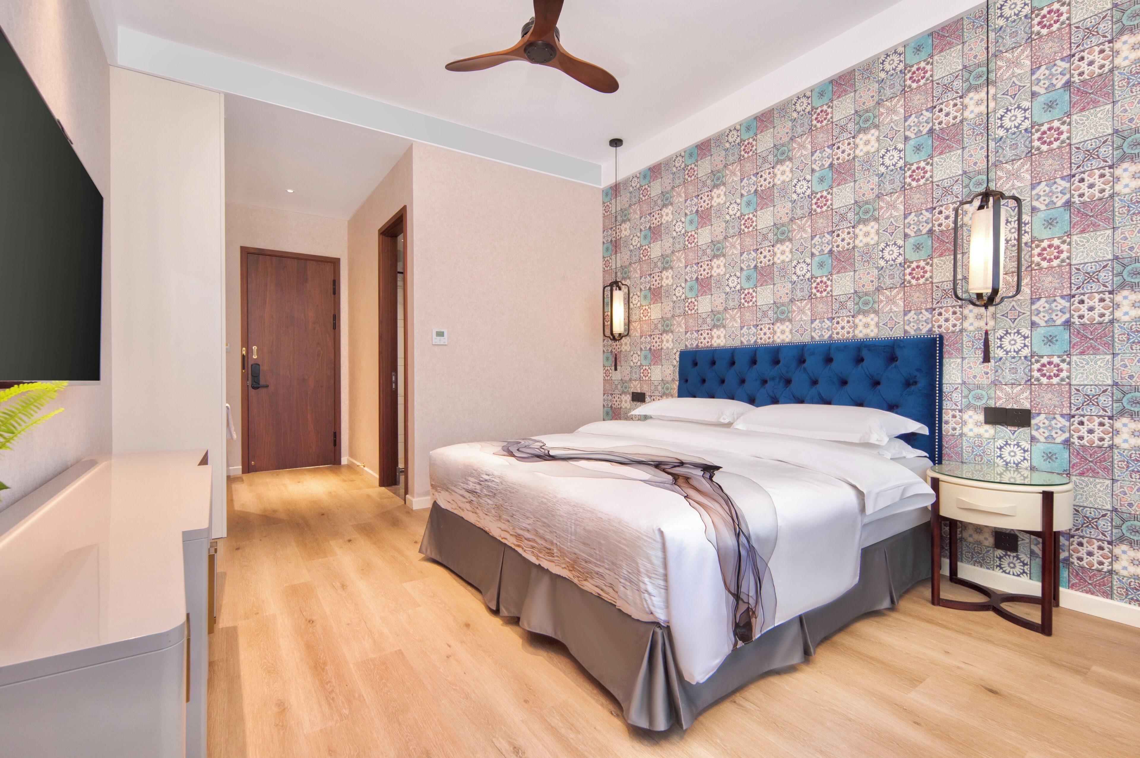 Un dormitorio con una cama, una mesita de noche, un ventilador de techo, un papel tapiz estampado y una puerta de madera.