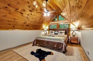 Ein Schlafzimmer mit Holzbalkendecke, einem Bett mit gemustertem Überwurf, einem Nachttisch mit Lampe und einem Fenster mit Blick auf Bäume.