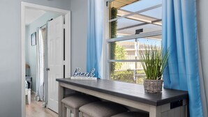 A hallway with a bench, a sign reading "Beach," and a window with blue curtains. - AMI Escape in 10 Min! Pool, Spa, Firepit & Game Room – Pet Friendly (Bradenton)