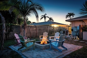 A backyard with a fire pit surrounded by Adirondack chairs, a wooden fence, and palm trees. - AMI Escape in 10 Min! Pool, Spa, Firepit & Game Room – Pet Friendly (Bradenton)