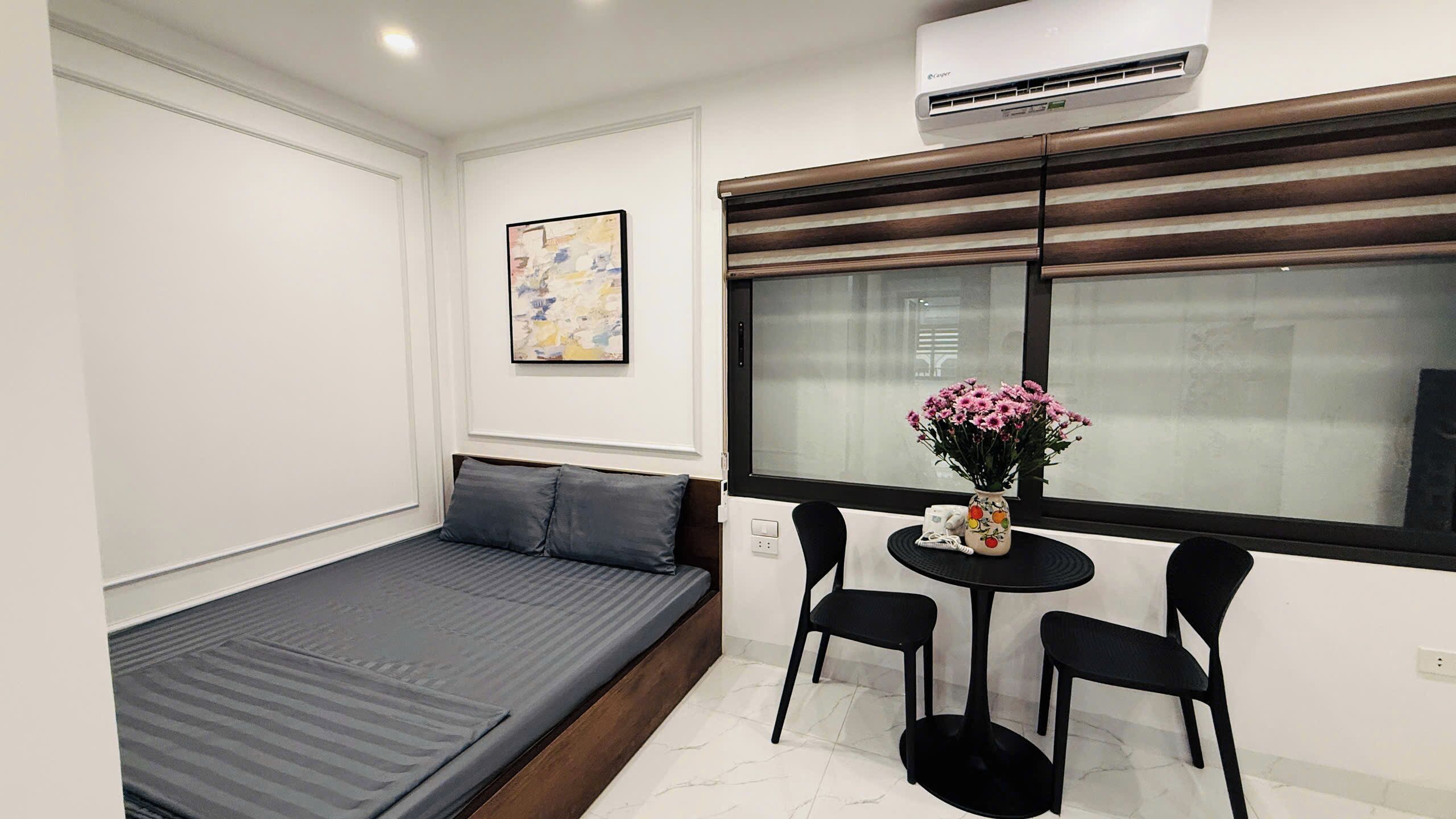 A compact room with a bed, a small table with chairs, a vase with flowers, and an air conditioning unit.