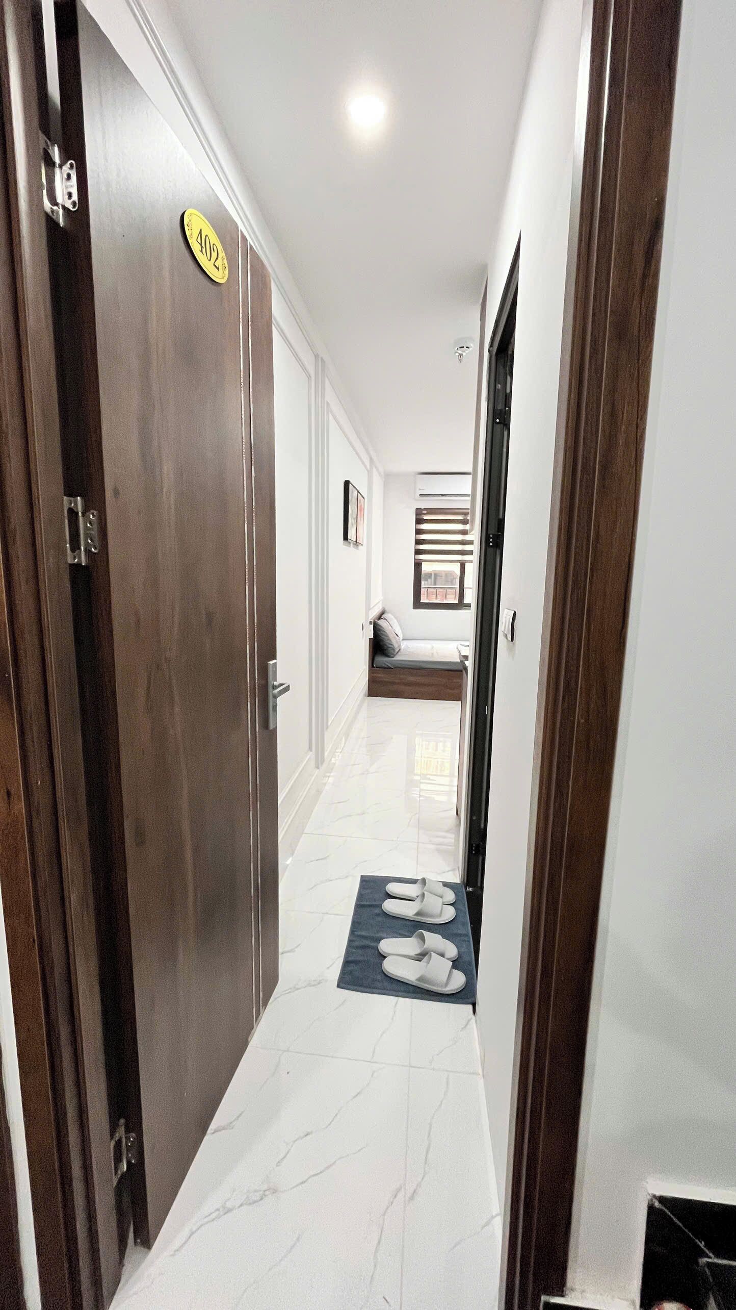 A hallway with a door marked '402', a bench, and a pair of slippers on a mat.