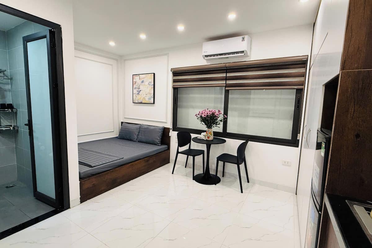 A compact living space with a sofa, a small table with chairs, a window with blinds, and an air conditioning unit.