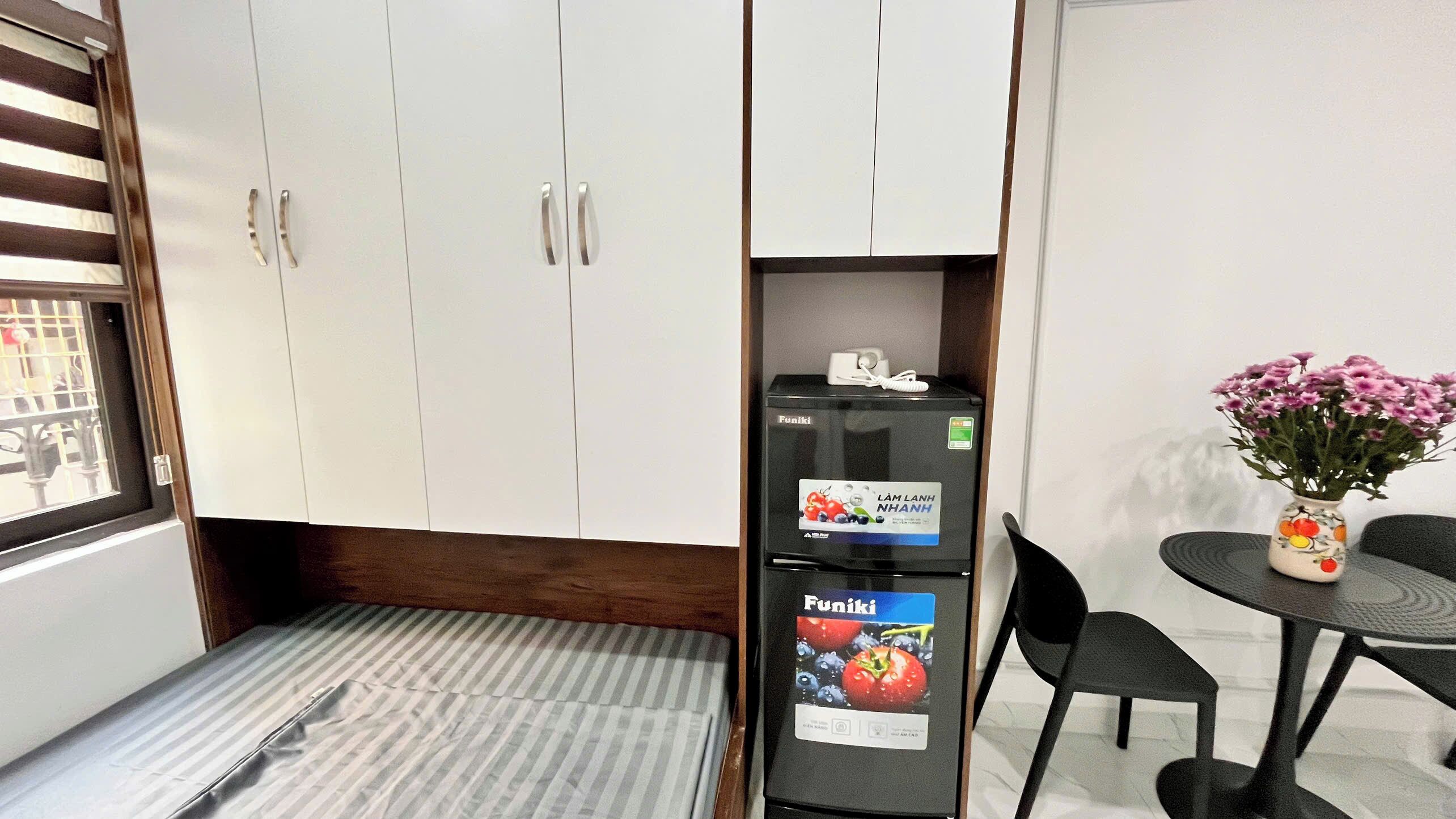 A compact room with a bed, a small table with chairs, a refrigerator, and a window with blinds.