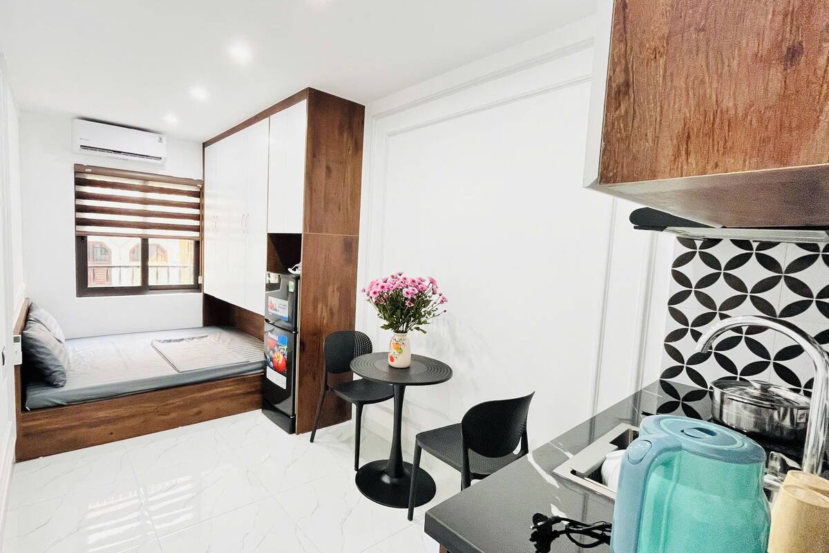 A compact hotel room with a bed, a small dining table, a chair, a sofa, a window with blinds, a wall-mounted air conditioner, and a decorative vase with flowers.