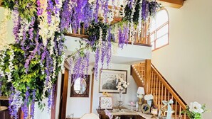 Een kamer met een houten trap, een eettafel met een kantonezen kleed en paars en witte bloemen die hangen.
