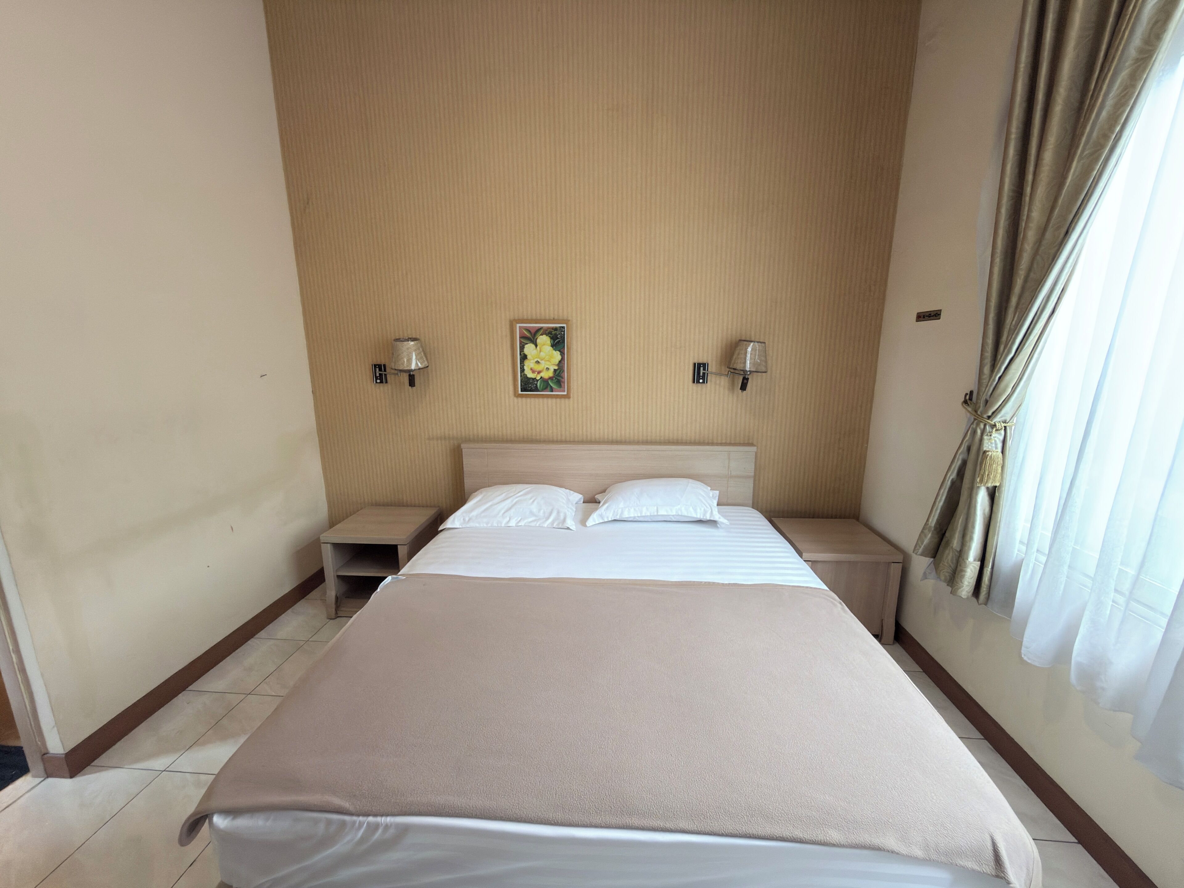 Una habitación de hotel con una cama grande, dos mesitas de noche, una lámpara de pared y un ventanal con cortinas.