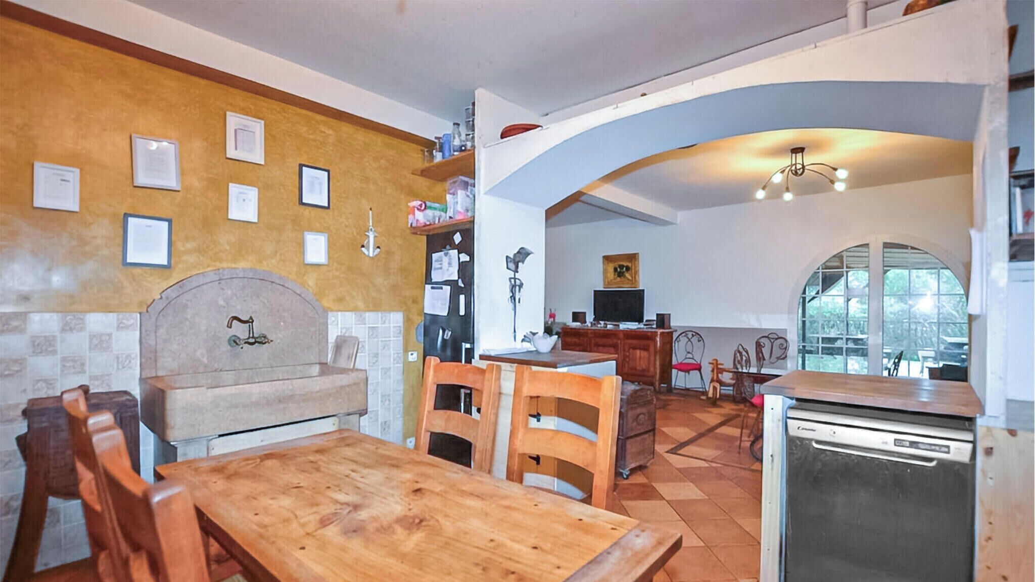 A kitchen with a wooden table, chairs, a sink, and a dishwasher. There is a tiled backsplash and a window with a view.