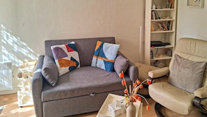 Un canapé gris agrémenté de coussins colorés, une table d’appoint blanche avec un vase et une boîte à mouchoirs, et un fauteuil beige.