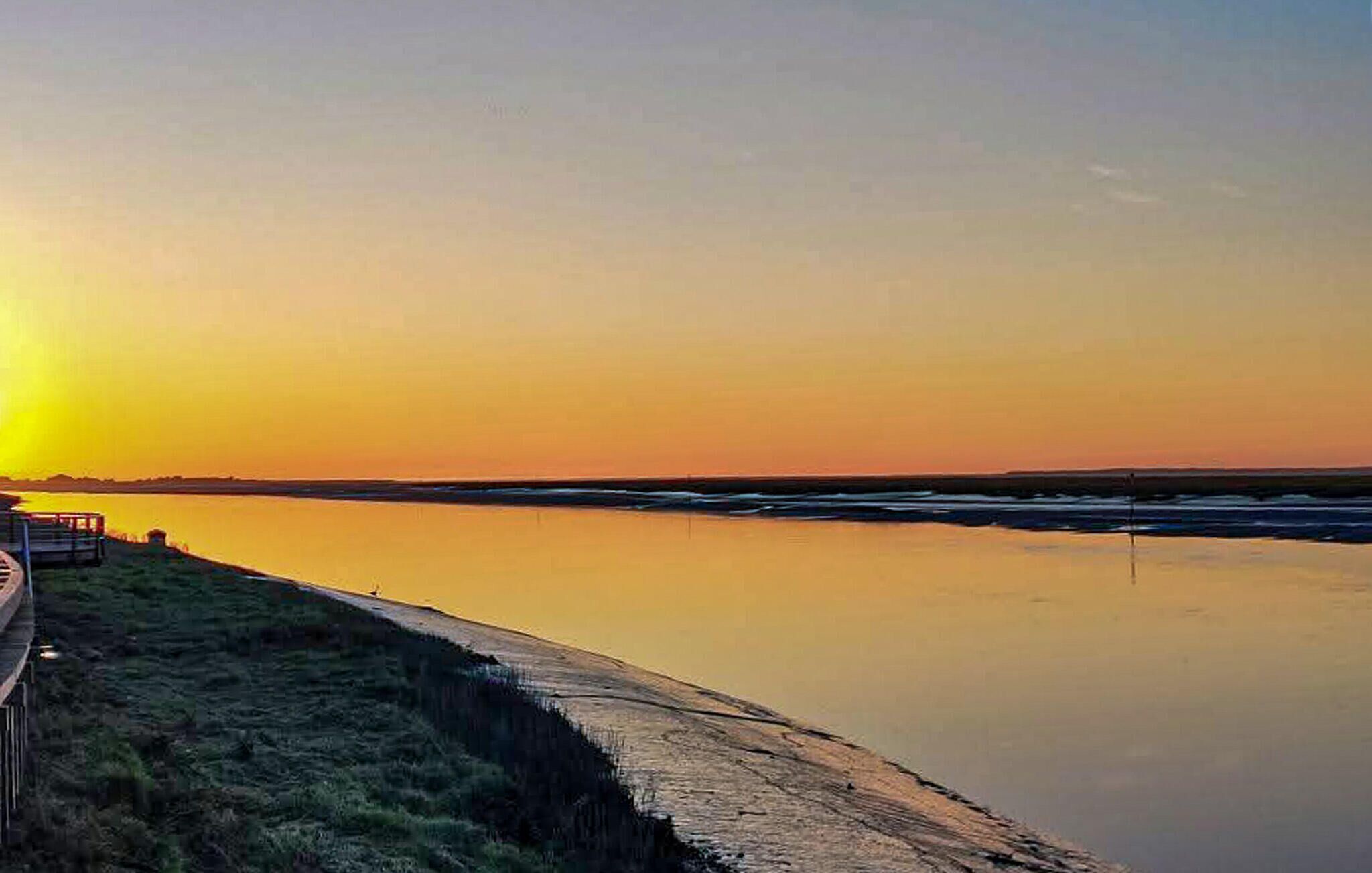 Solnedgang over rolige vand med en træbro og græsareal.