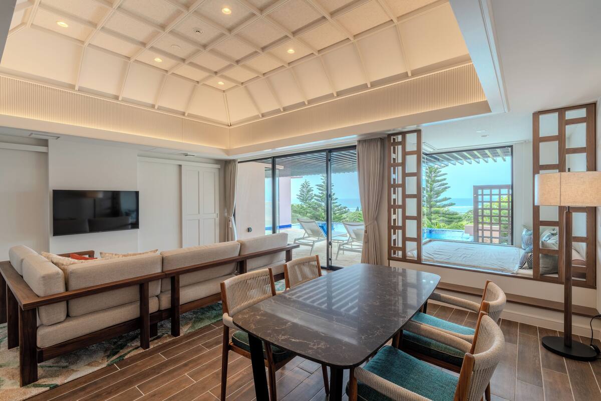 Un vaste espace de vie comprenant un canapé, une table à manger et des chaises, un téléviseur à écran plat fixé au mur et de grandes fenêtres offrant une vue sur l’extérieur.