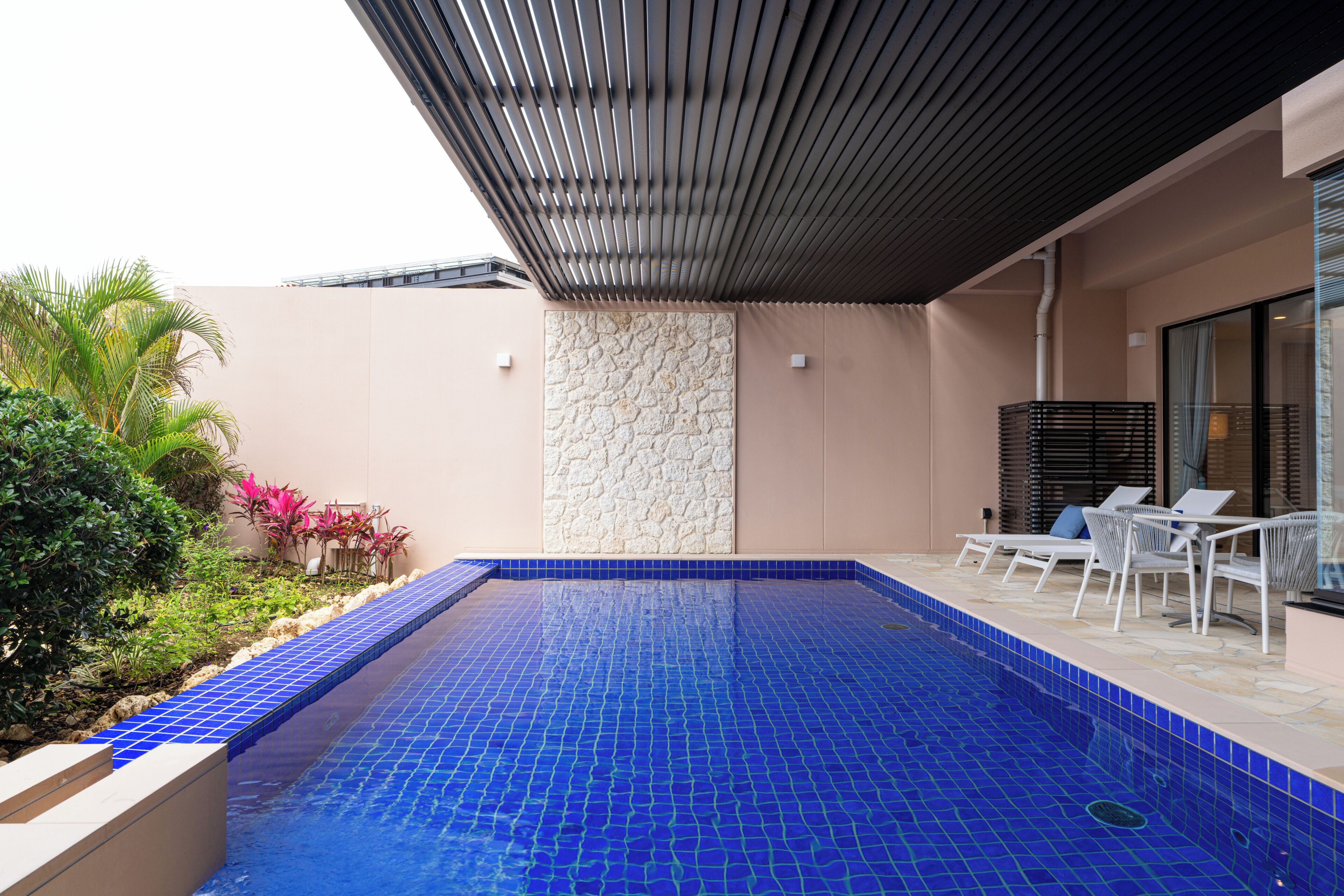 Ein Swimmingpool mit blauen Fliesen, umgeben von Pflanzen und einem überdachten Sitzbereich.