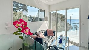 Een moderne woonkamer met een glazen salontafel, een bank met kussens, een vaas met bloemen en uitzicht op de zee door grote ramen.