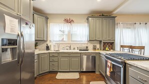 Una cocina con gabinetes grises, electrodomésticos de acero inoxidable, horno empotrado y una ventana con cortinas blancas.