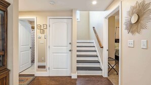 Un pasillo con suelo de madera, una puerta blanca, una escalera con escalones negros y un espejo fijado a la pared.