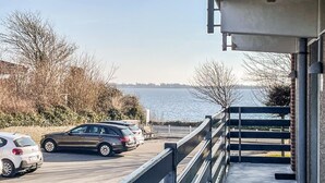 Un balcon avec vue sur l’eau, un espace de stationnement avec des voitures et une clôture blanche.