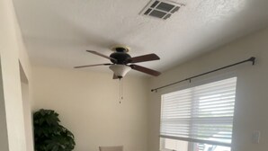Ein Essbereich mit Tisch, Stühlen, Deckenventilator, Fenster mit Jalousien und einer Topfpflanze.