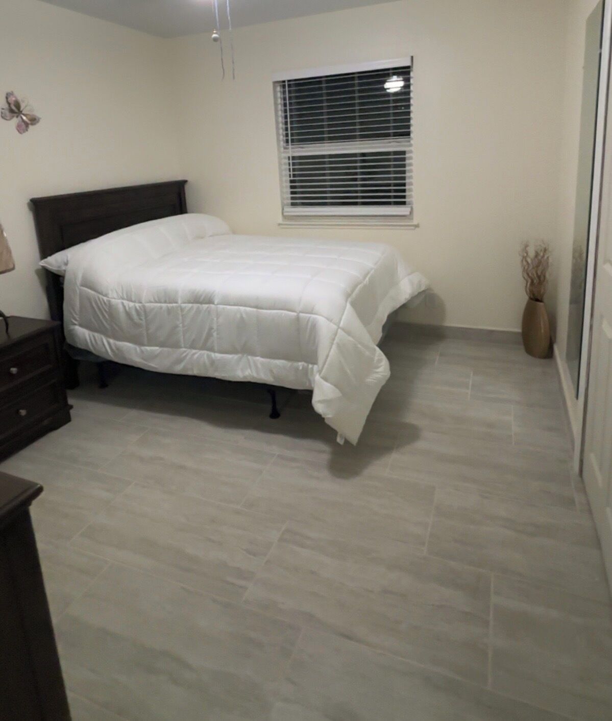Une chambre à coucher comprenant un lit, une commode, une fenêtre avec des stores et un vase contenant des plantes séchées.
