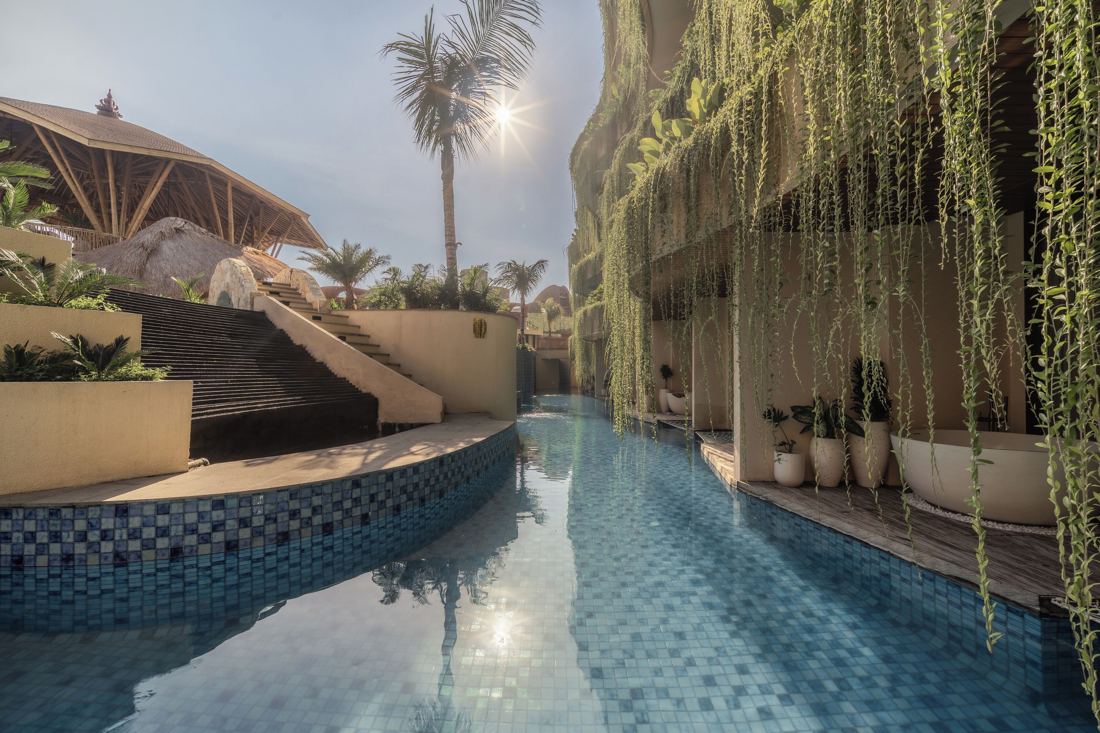 Una piscina con bordes de azulejos, rodeada por un edificio con techo de paja y una escalera que sube a un nivel superior.
