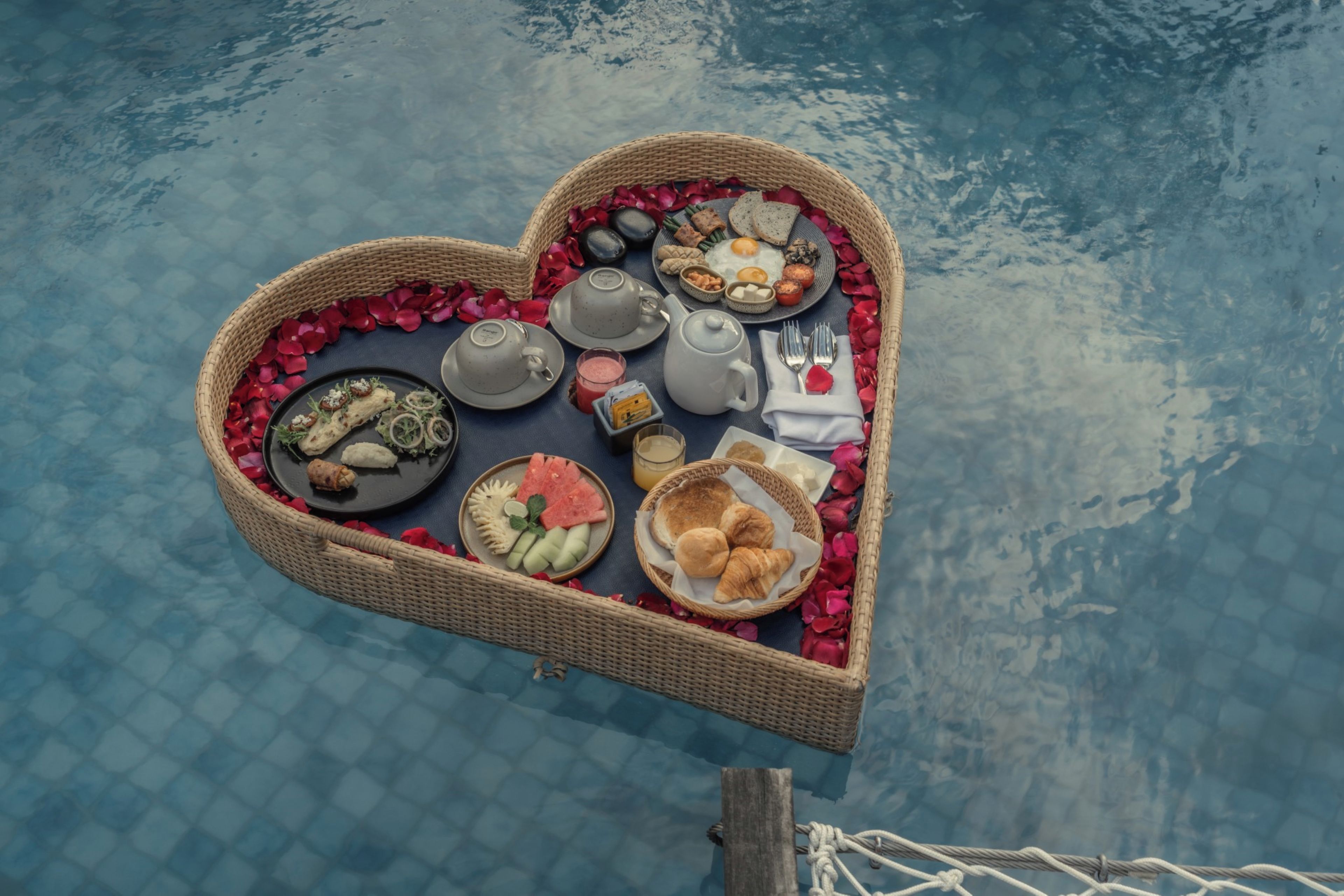 A heart-shaped table with a variety of food items and a teapot, placed on a dock over a body of water.
