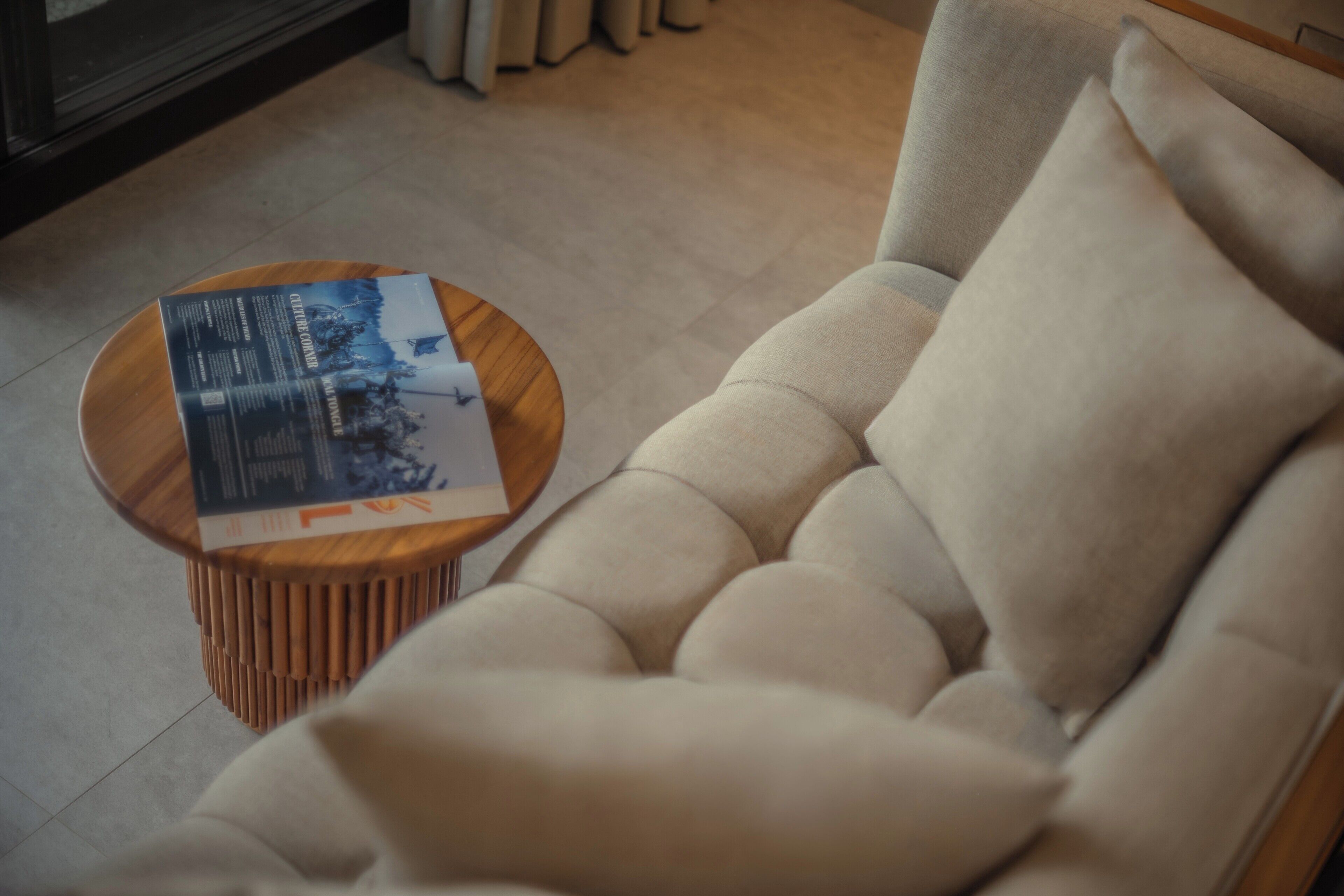 Una sala de estar moderna con un sofá beige, una mesa de centro redonda de madera con una revista, y un suelo de baldosas.