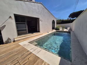 Ein Swimmingpool mit Holzterrasse, einer Schiebetür und einem kleinen Sitzbereich mit zwei Stühlen.