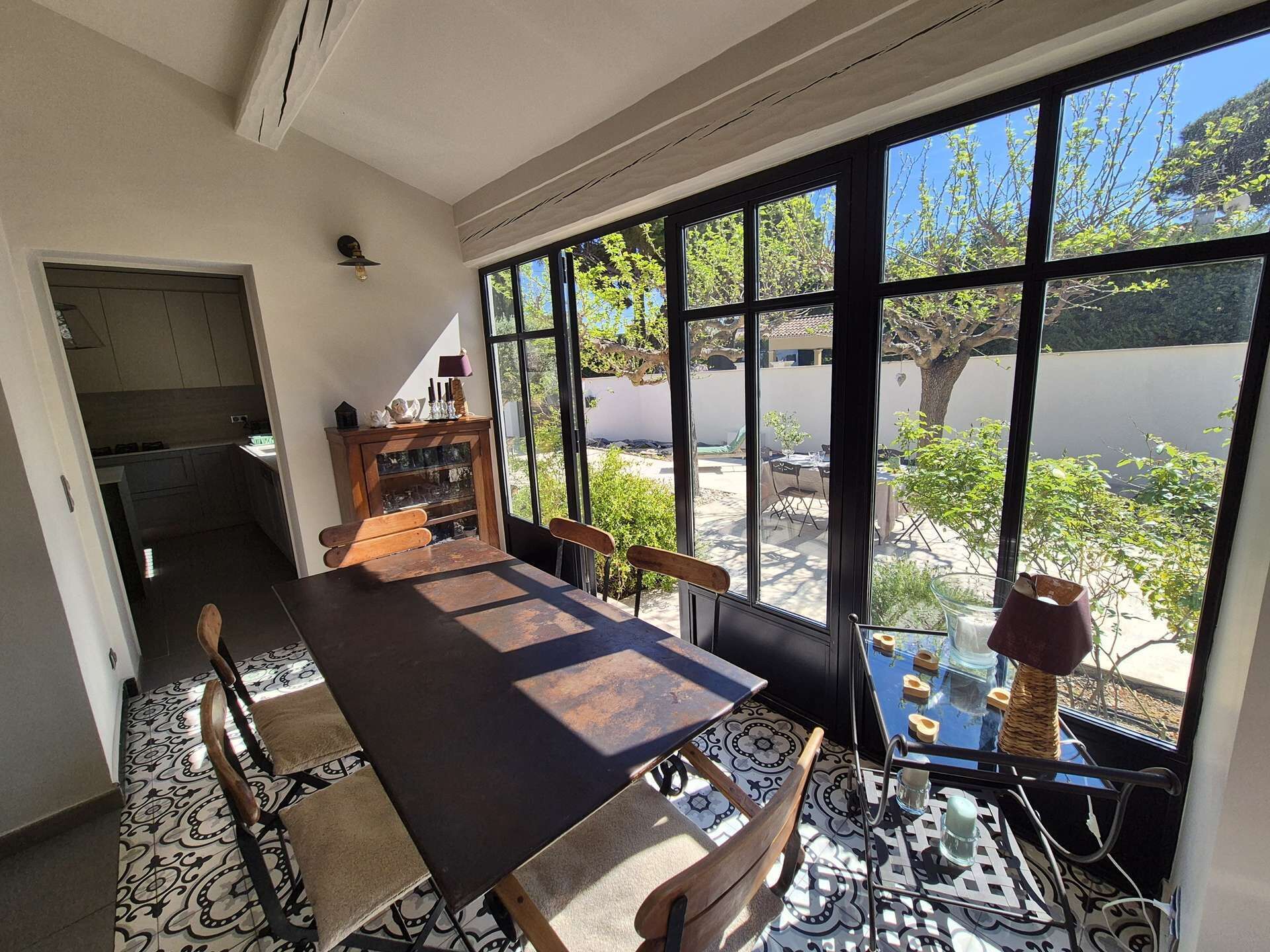 A dining area with a large table, chairs, a small table with a vase, and a view of the outdoors through large windows.