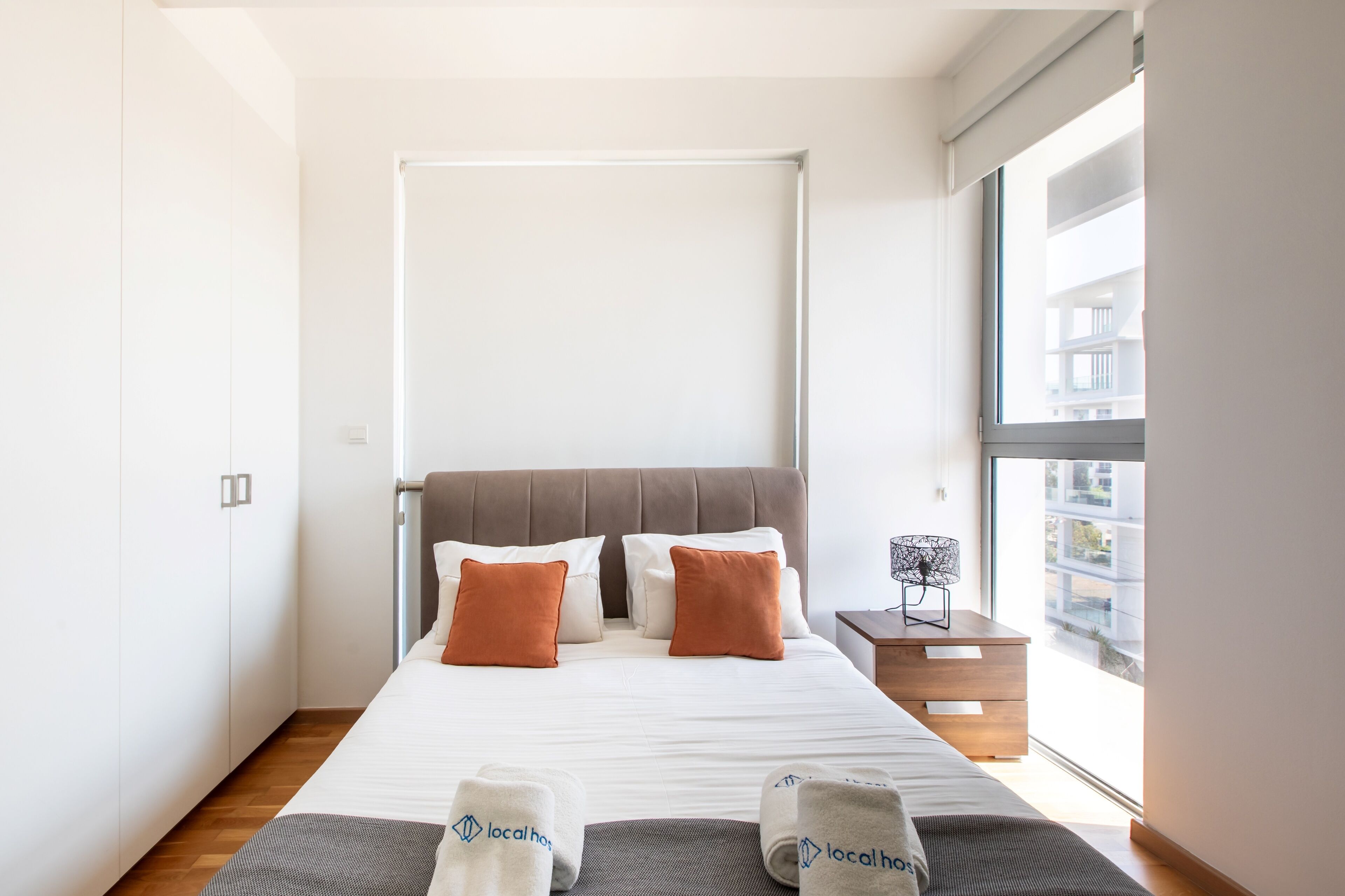 Habitación de hotel con una cama grande, una ventana con vistas a edificios, una mesita de noche con lámpara y toallas con la marca 'local host'.