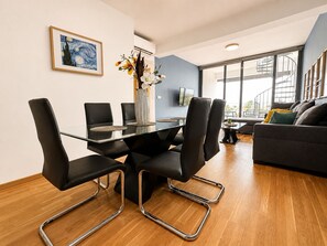 A modern living room with a glass dining table, black chairs, a sofa, and a large window.