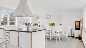 Moderni keittiö, jossa on valkoiset kaapit, keskellä sijaitseva keittiösaareke ja ruokailutila, jossa on valkoiset tuolit.