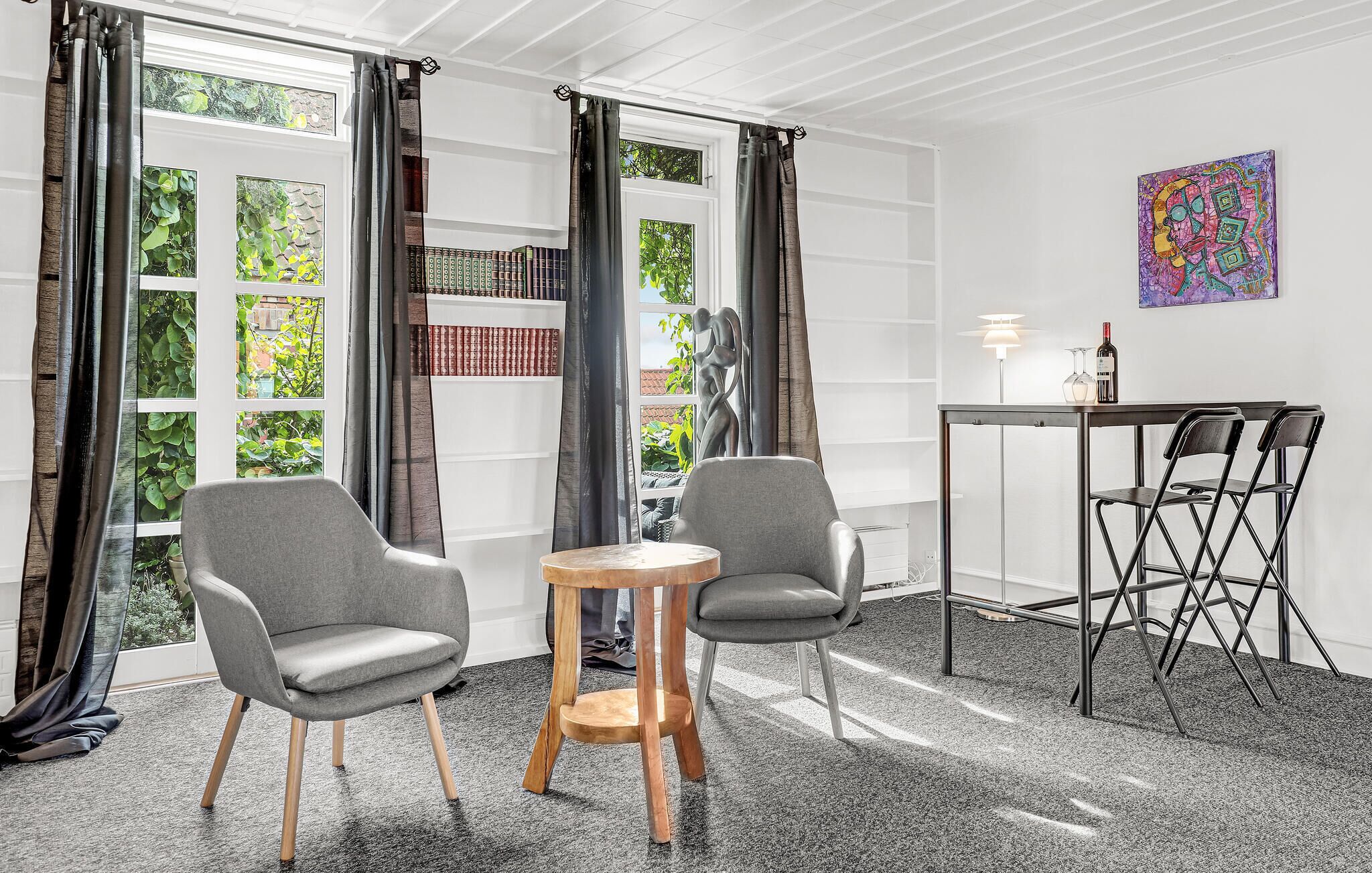 Una habitación con dos sillas grises, una mesita redonda pequeña, una mesa plegable de metal negro con sillas, una lámpara y una estantería con libros.