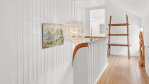 Käytävän seinät ovat valkoiset ja niissä on paneelit, lattia on puuta, seinällä on taulu, siellä on ainutlaatuinen valaisin ja puinen tikapuu.