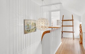 A hallway with white paneled walls, a wooden floor, a painting, a unique light fixture, and a wooden ladder.