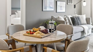 A round dining table with a coffee pot, a fruit platter, and a cup of coffee. There are two chairs around the table. The room has a grey couch, a white armchair, and a wall with framed pictures.